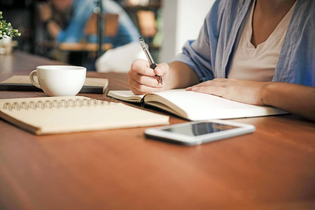 Student writes a creative story in a notebook with a cup of coffee and a phone nearby.