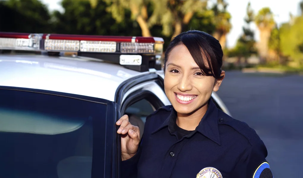 A police officer leaning against the side of a patrol car, smiling, and looking directly at the camera.