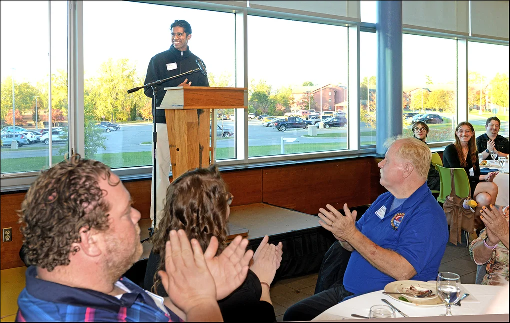A young man at a podium with attendees around tables clapping