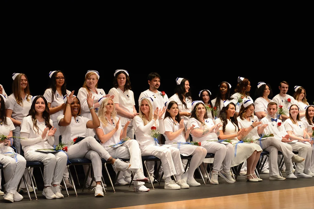 Two rows of nurses in white uniforms, one seated and one standing, clapping at ceremony