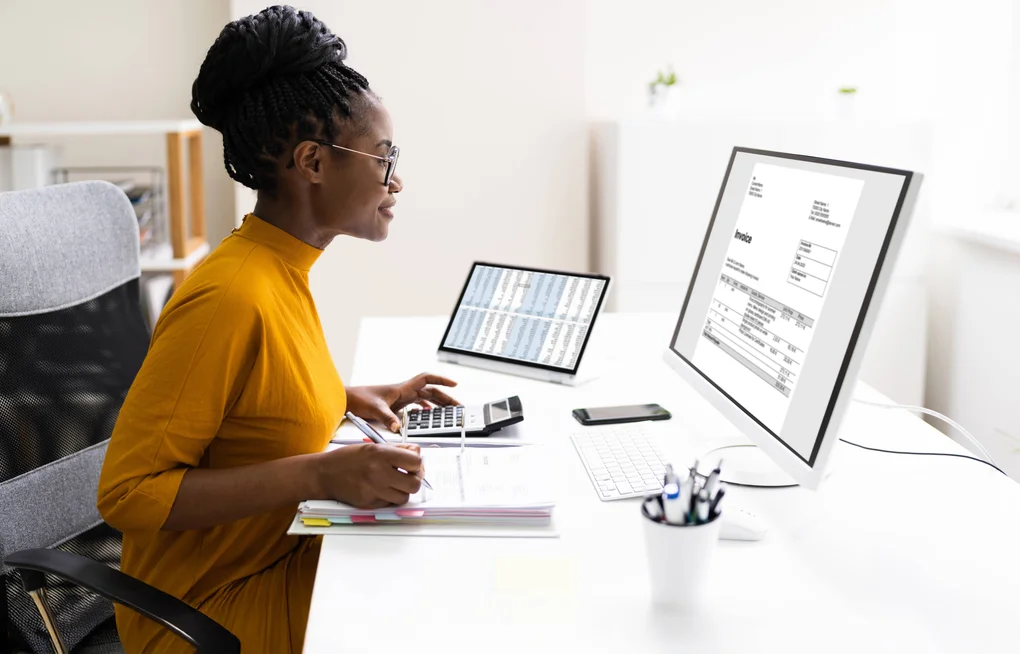 An accountant sits at her desk reviewing invoices on a screen while inputting figures into a calculator.