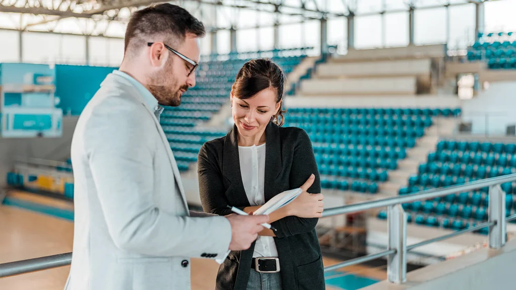 Two Sports Studies students stand in the middle of an empty gymnasium reviewing a playbook together.