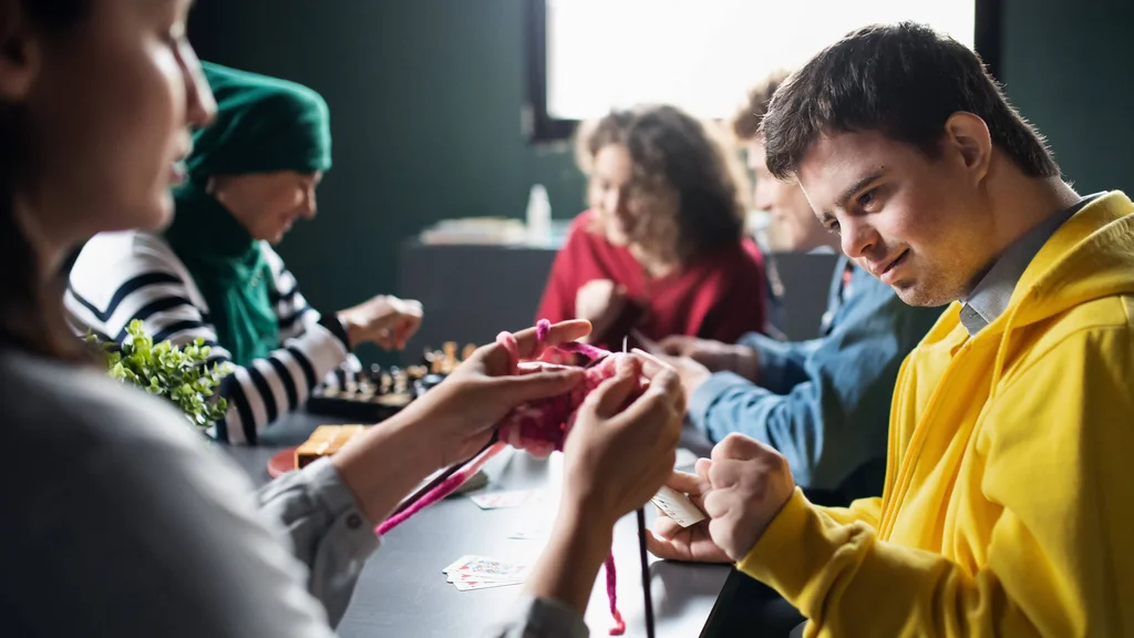 Direct Support Professionals and their clients gathered around a table, knitting, and playing chess.