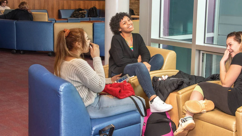 A group of three students relax in a campus lounge, smiling and laughing.
