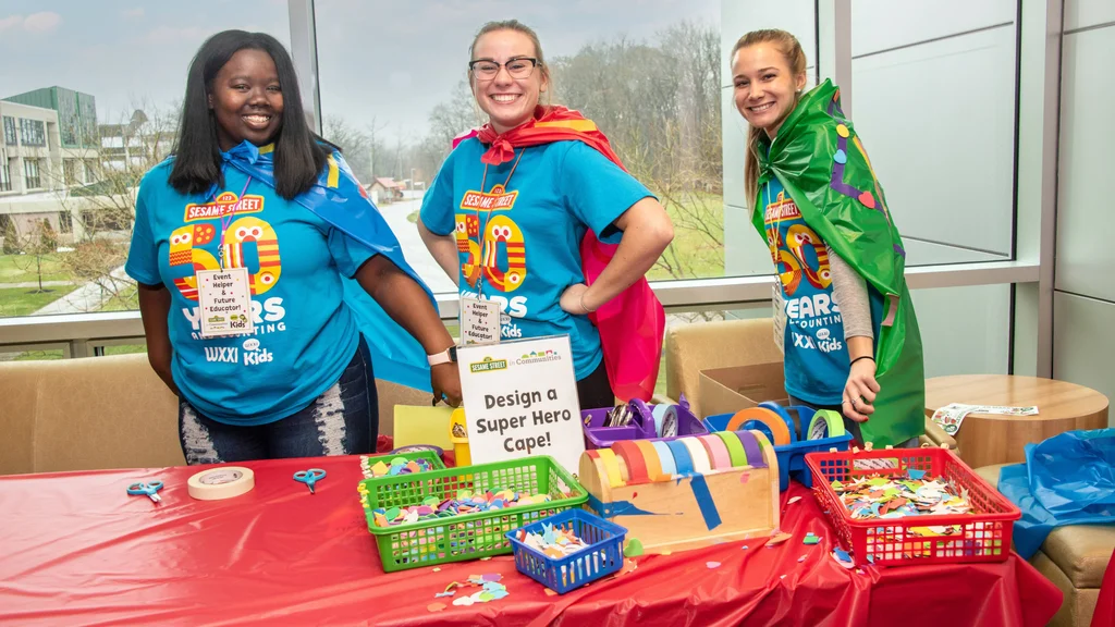 Three smiling college students. Each wears a shirt celebrating the 50th anniversary of Sesame Street and a colorful, homemade cape.