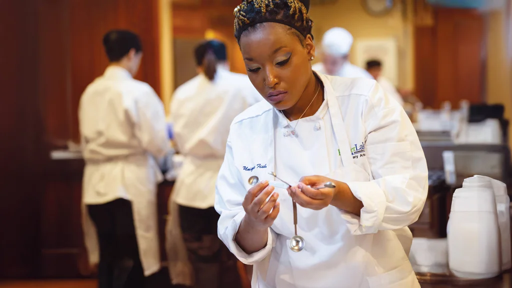 A student in a white chef's jacket and apron. She inspects a set of measuring spoons as other students prepare food in the background.