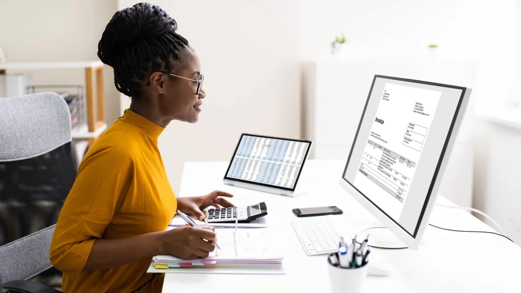 An accountant sits at her desk reviewing invoices on a screen while inputting figures into a calculator.