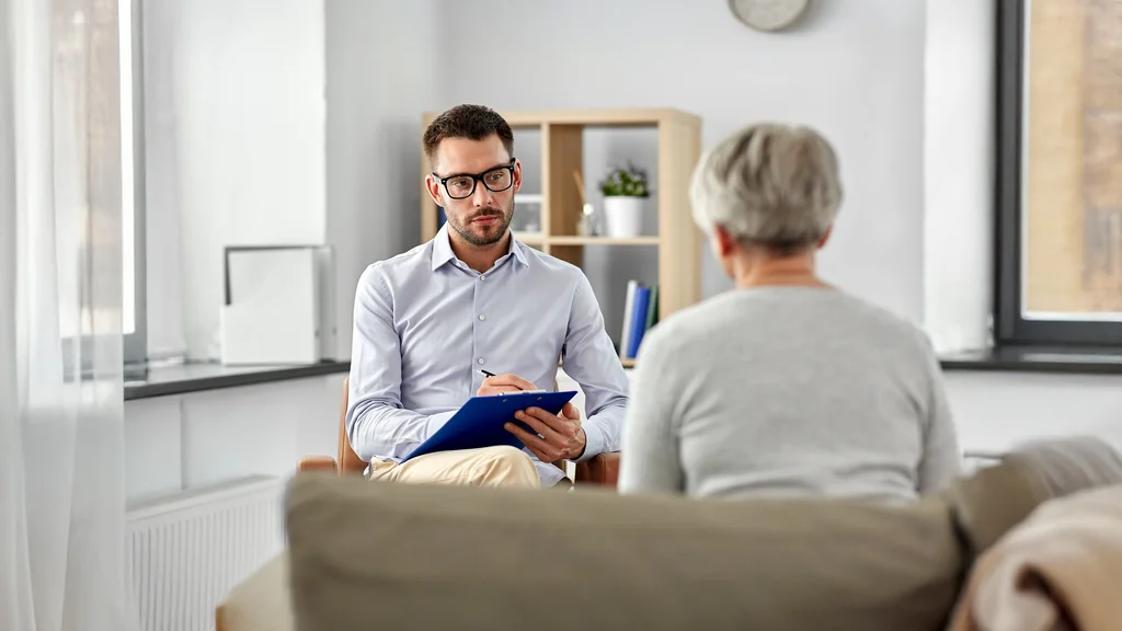 A counselor listens to a patient speaking. The counselor keeps notes of the conversation.