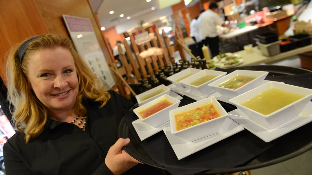A server smiles at the camera while holding a food tray loaded with several bowls of soup prepared by FLCC Culinary Arts students.