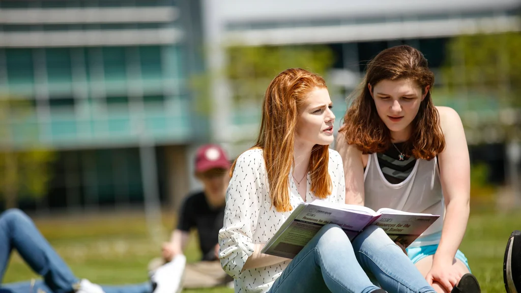 Two young women read from a college textbook together while lounging in the grass on a bright and sunny campus lawn.