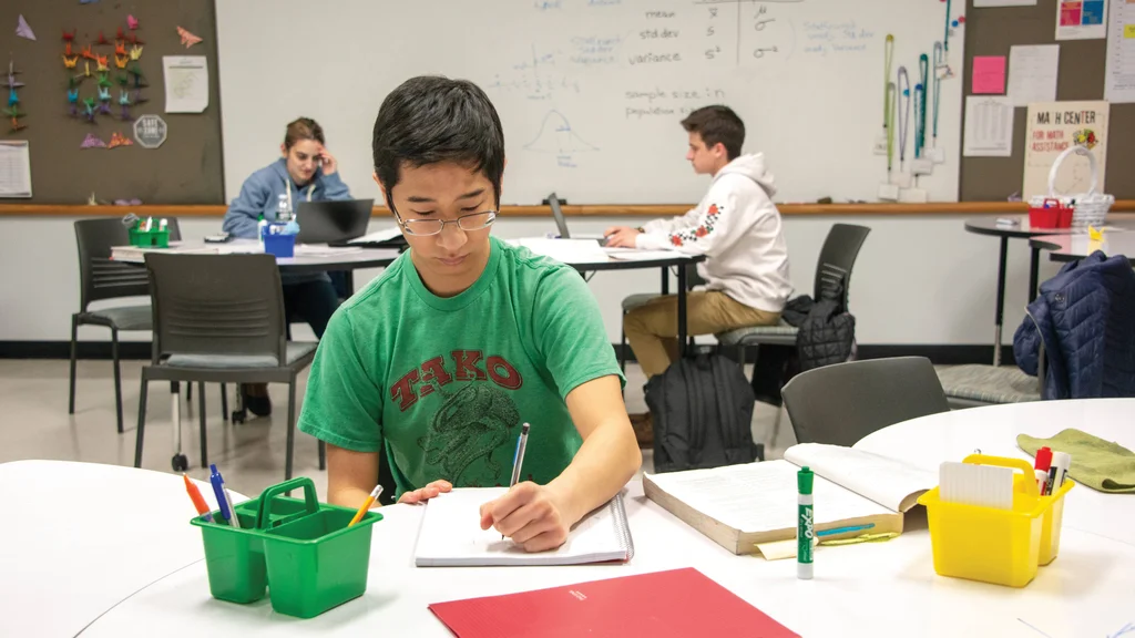 Three people studying in the FLCC Math Center. One student writes in a notebook while two others work on their laptops at nearby tables.