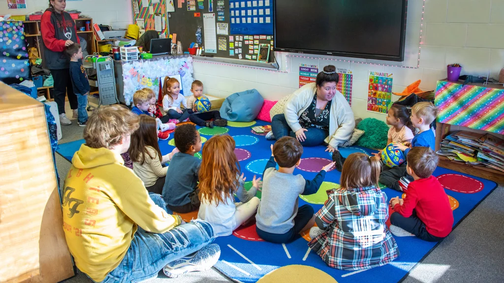 Several children sitting in a circle on a brightly colored carpet, engaging with the teacher during a large group activity.
