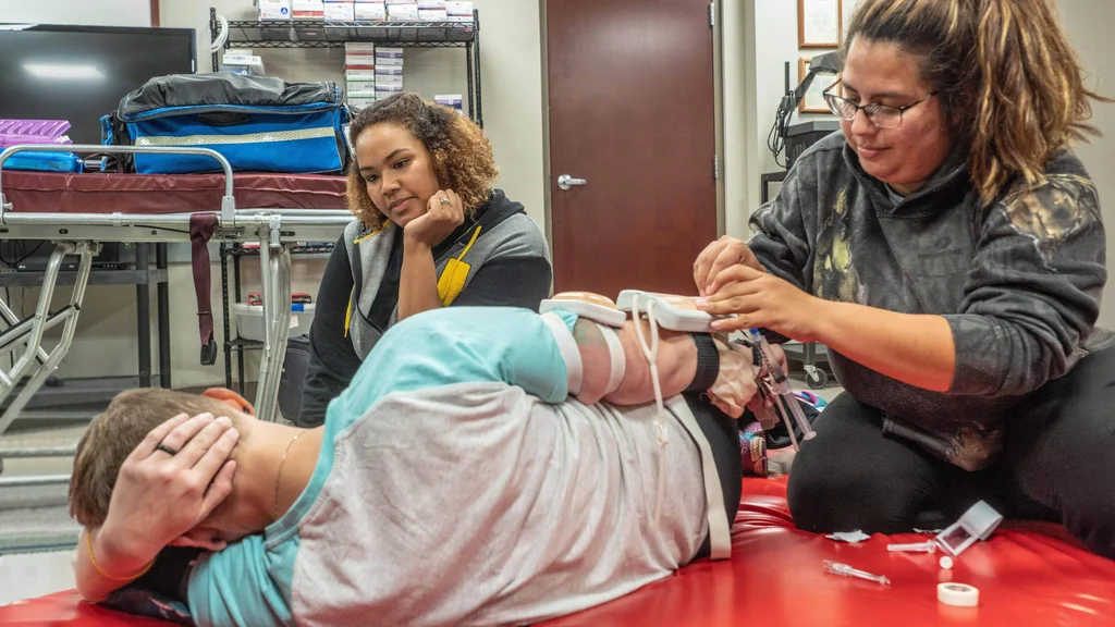Two EMT students practice securing medical monitoring devices to a third student's arm. The volunteer patient is lying on his side as though injured.