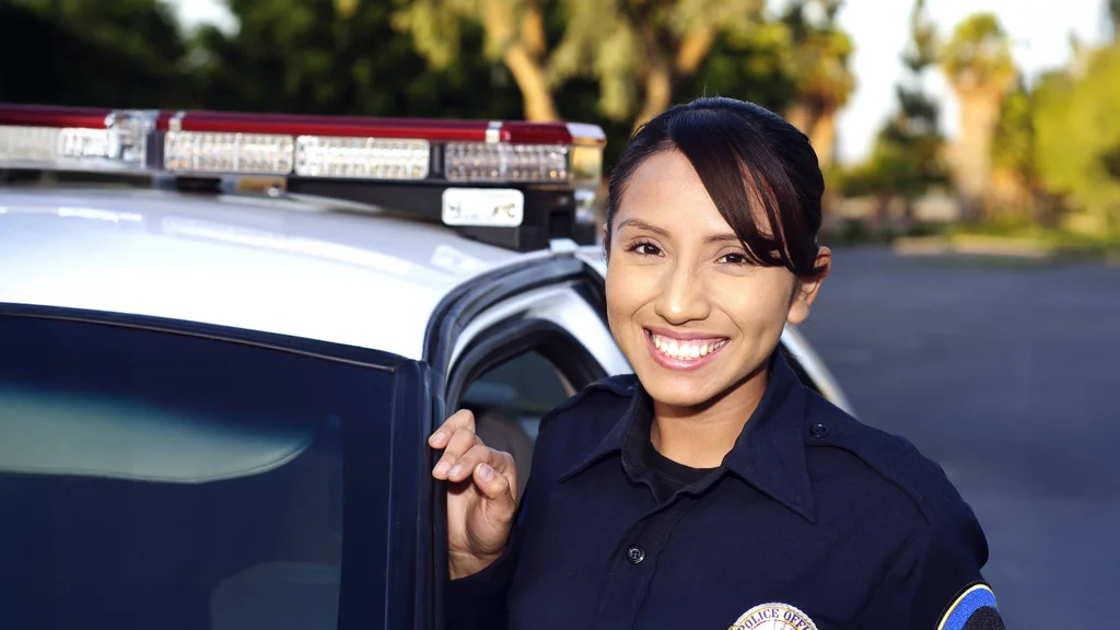 A police officer leaning against the side of a patrol car, smiling, and looking directly at the camera.