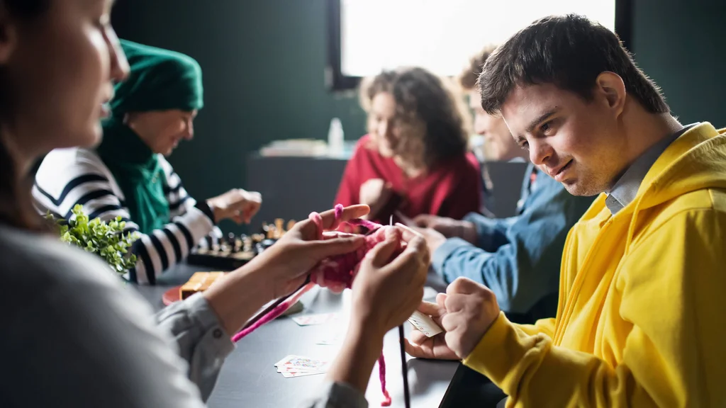 Direct Support Professionals and their clients gathered around a table, knitting, and playing chess.