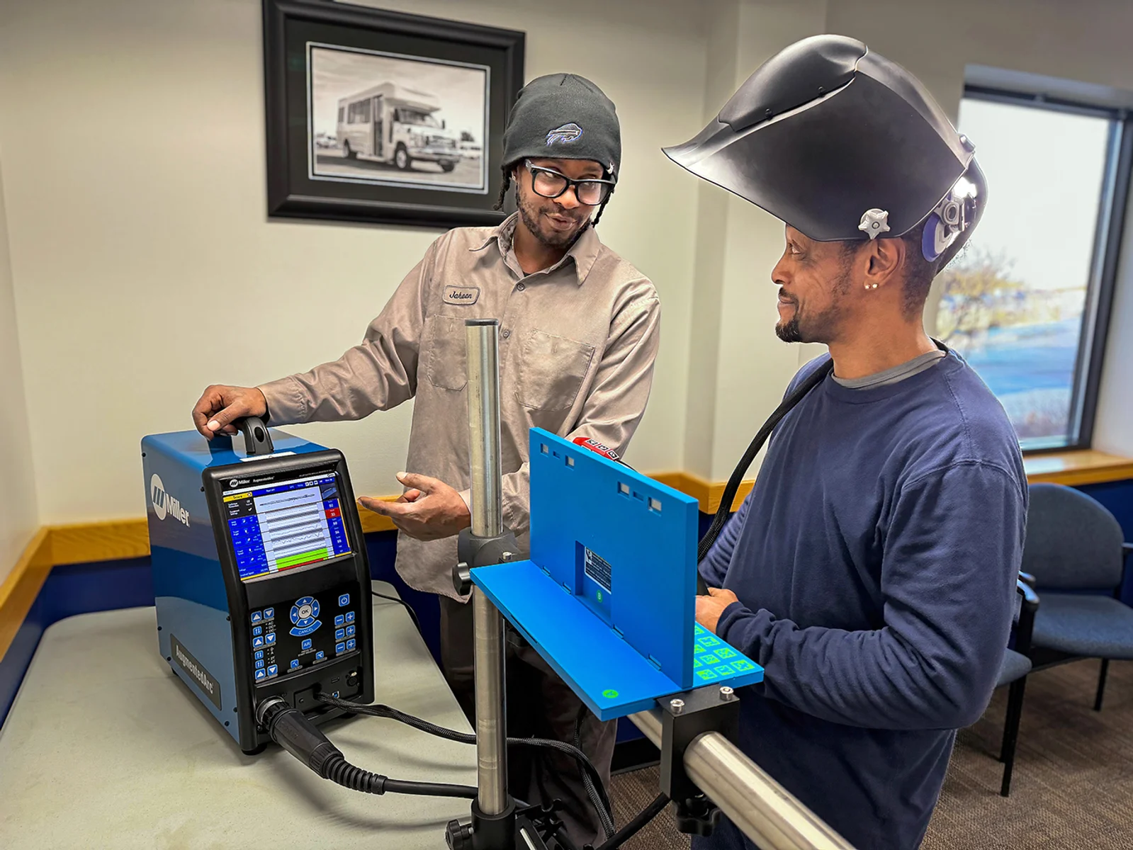 Instructor points to student using welding simulator, student has lifted up his welding helmet