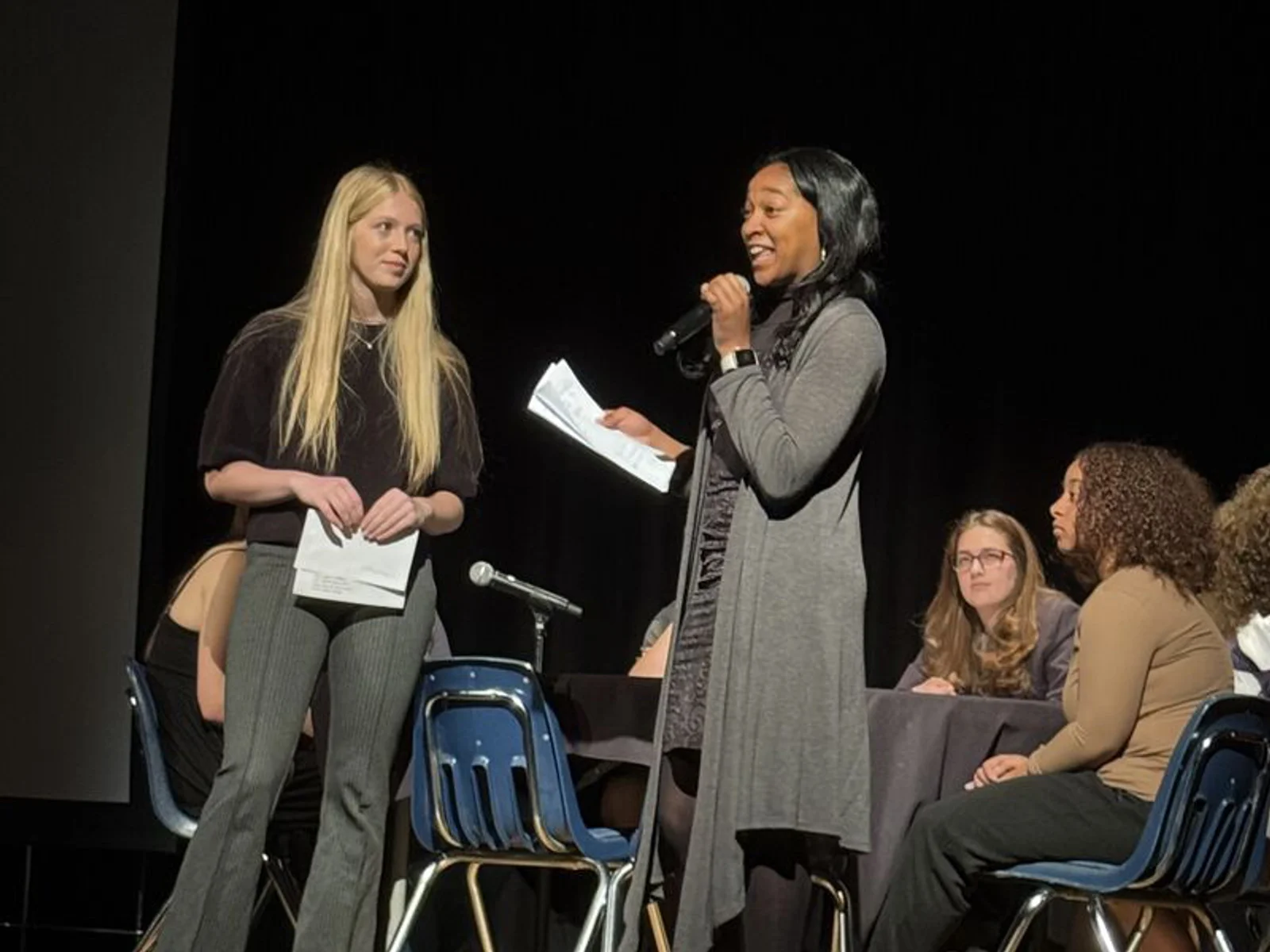 Two students standing on a stage, one speaking into a microphone, with other students seated behind them