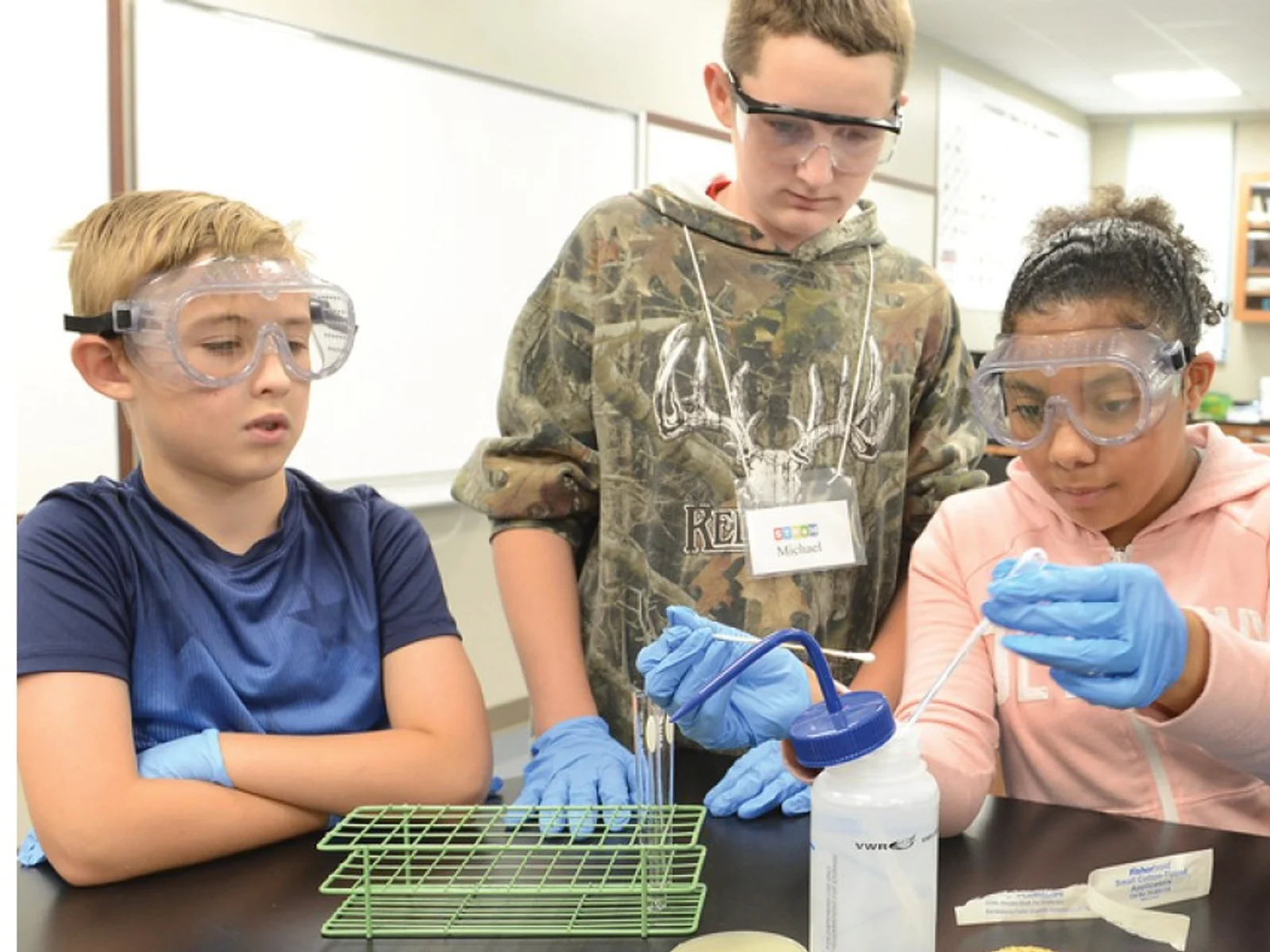 Three students wearing eye protection stand around a lab table as a fourth student uses instruments