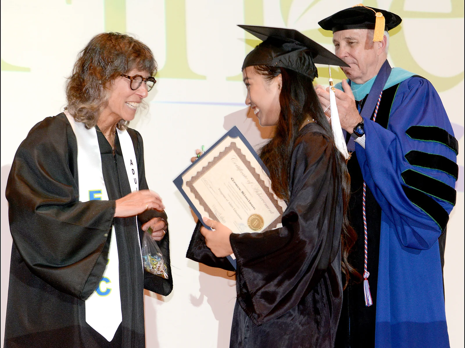 A woman in black cap and gown shakes hands with another women in regalia as a man in academic regalia looks on