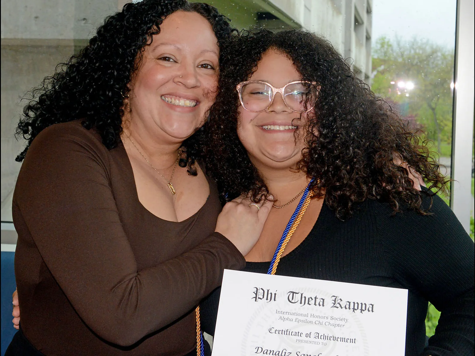 Two women lean toward one another and pose, smiling, as one holds an honor society certificate
