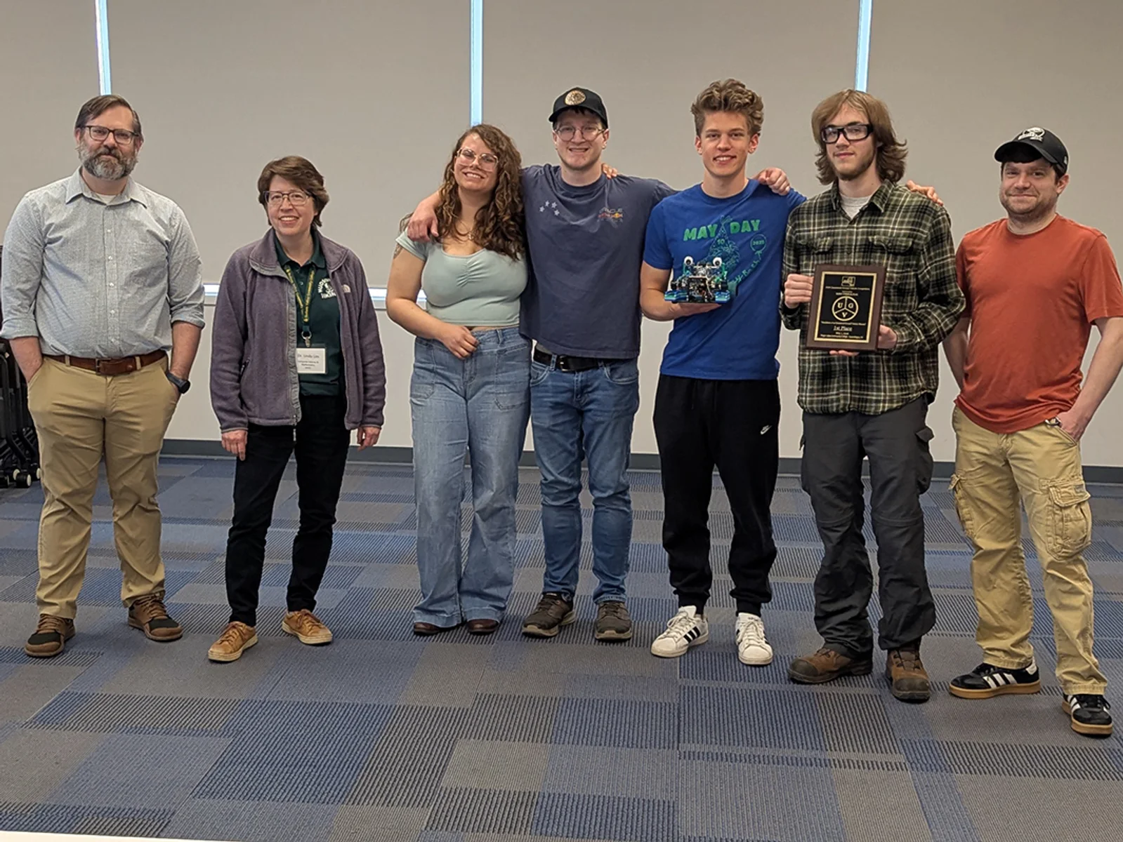 Seven people standing in a row, one holding a plaque