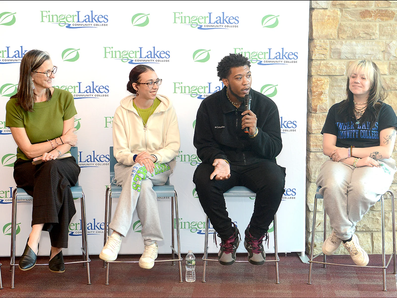Four people sitting in a row on stools as one speaks into a microphone