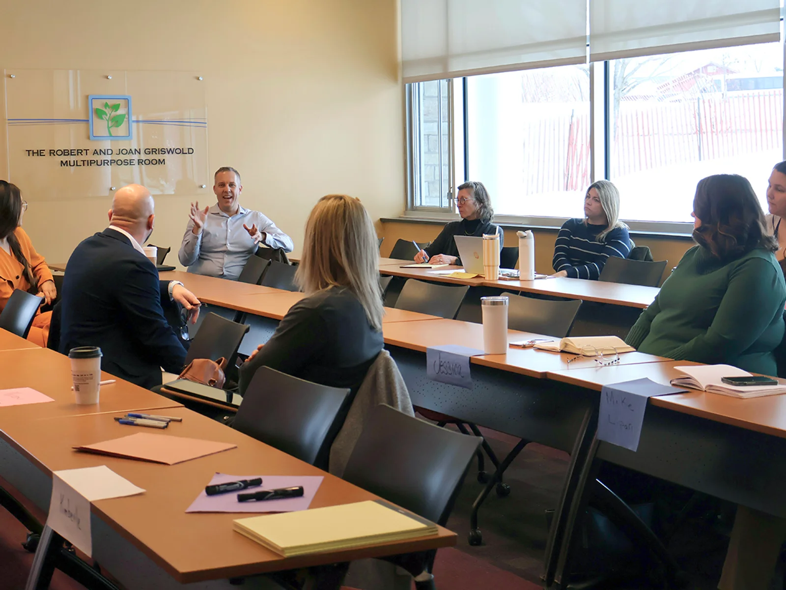 Workshop participants seated at three rows of tables, looking toward a man speaking at one end