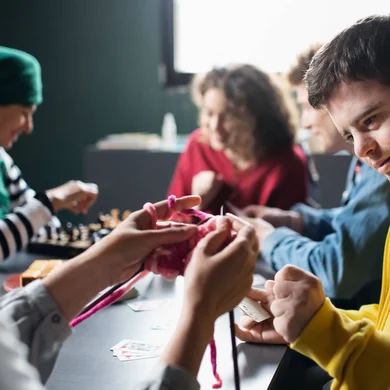 Direct Support Professionals and their clients gathered around a table, knitting, and playing chess.