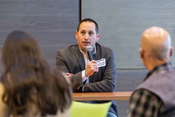 Man, seated, speaking into microphone before audience