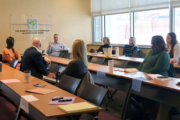 Workshop participants seated at three rows of tables, looking toward a man speaking at one end