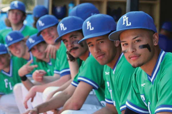 The men's baseball team sit in a dugout, ready for the game to start.
