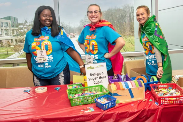 Three smiling college students. Each wears a shirt celebrating the 50th anniversary of Sesame Street and a colorful, homemade cape.