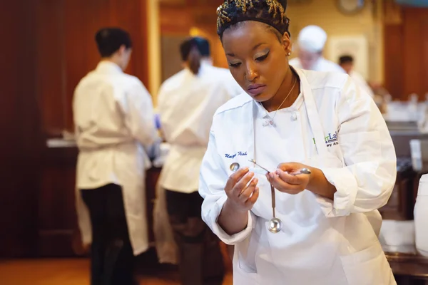 A student in a white chef's jacket and apron. She inspects a set of measuring spoons as other students prepare food in the background.
