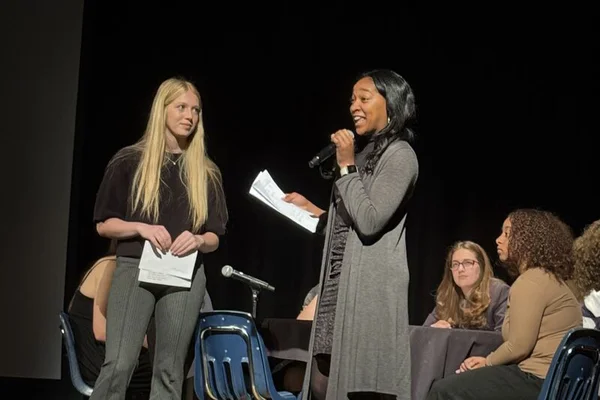 Two students standing on a stage, one speaking into a microphone, with other students seated behind them