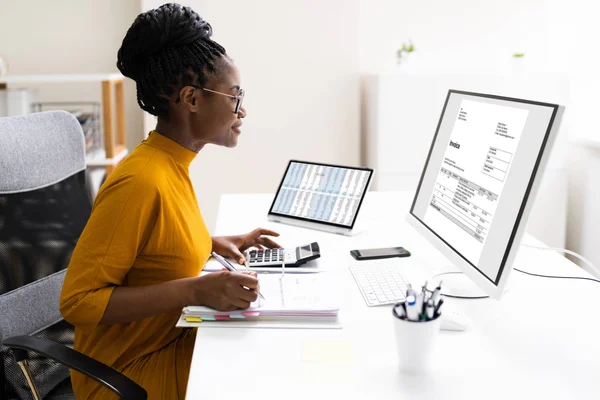 An accountant sits at her desk reviewing invoices on a screen while inputting figures into a calculator.