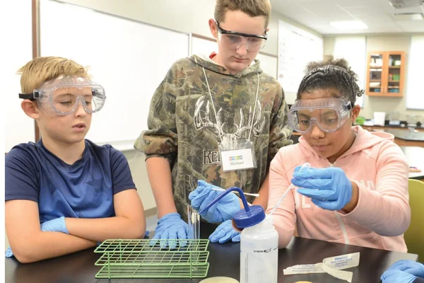 Three students wearing eye protection stand around a lab table as a fourth student uses instruments