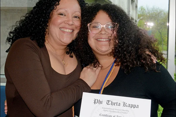 Two women lean toward one another and pose, smiling, as one holds an honor society certificate