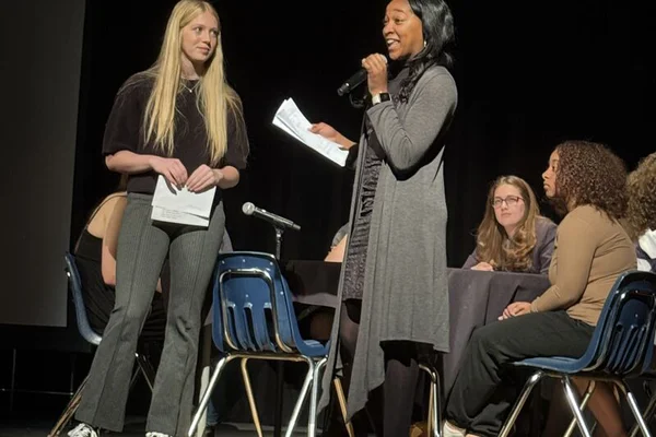 Two students standing on a stage, one speaking into a microphone, with other students seated behind them