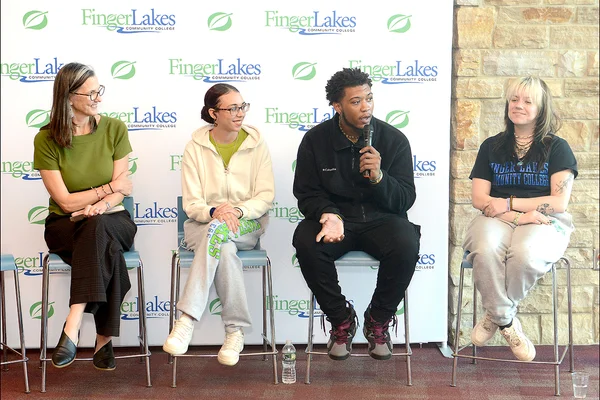 Four people sitting in a row on stools as one speaks into a microphone