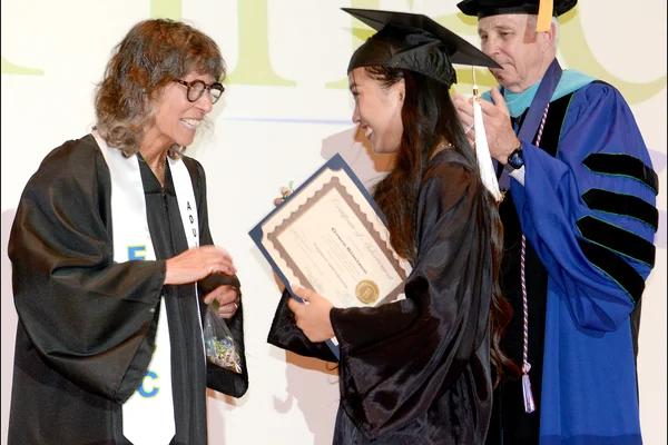 A woman in black cap and gown shakes hands with another women in regalia as a man in academic regalia looks on