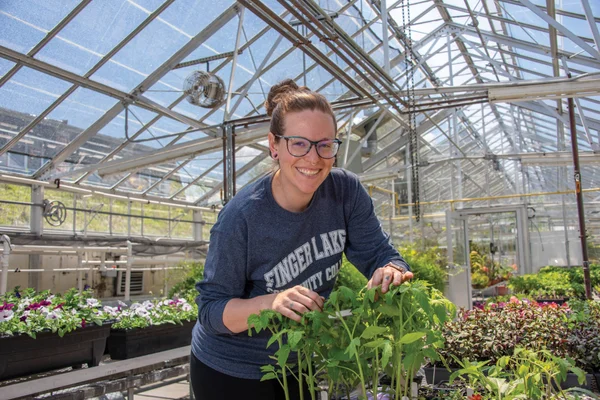 A young woman tending to her plants in the Finger Lakes Community College greenhouse pauses to smile at the camera.