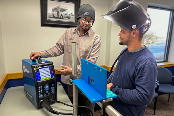 Instructor points to student using welding simulator, student has lifted up his welding helmet