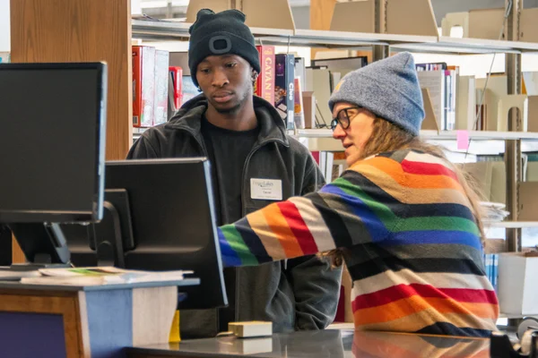 Two employees in of the Library work together at a computer near the reference section.