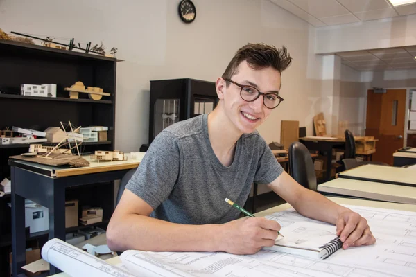 An architecture student works at a drawing table on blueprints at FLCC's Victor Campus Center. 3D models of buildings and a 3D printer are in the background.