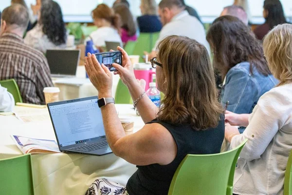 Woman at table with laptop taking photo with phone