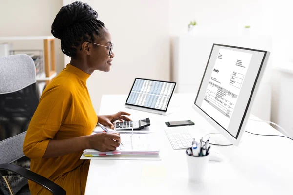 An accountant sits at her desk reviewing invoices on a screen while inputting figures into a calculator.