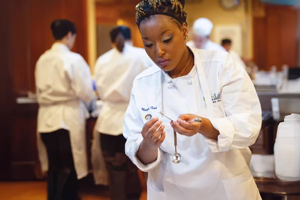 A student in a white chef's jacket and apron. She inspects a set of measuring spoons as other students prepare food in the background.