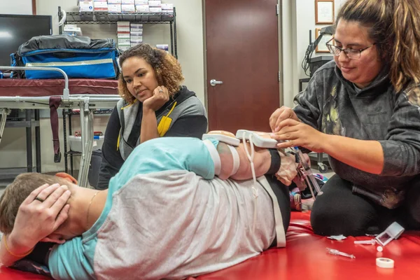 Two EMT students practice securing medical monitoring devices to a third student's arm. The volunteer patient is lying on his side as though injured.