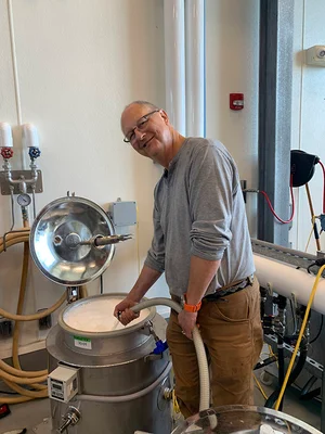 An older male student looks forward and smiles as he holds a hose into a stainless steel vat in the viticulture lab