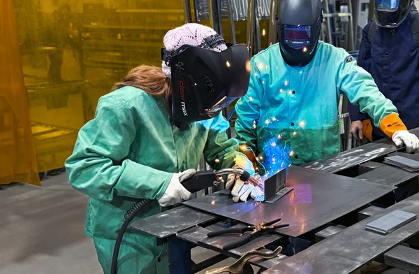 Young woman welding in helmet and protective smock while another worker watches